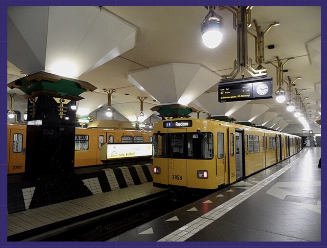 Foto eines gelben Berliner U-Bahn-Zugs (Baureihe F, Nummer 2858) der Linie U7 Richtung Rudow, stehend an einem Bahnsteig. Der Bahnhof hat eine weiße Gewölbedecke, schwarze Stützpfeiler mit Art-déco-Ornamenten und charakteristisches schwarz-weißes Bodendesign.